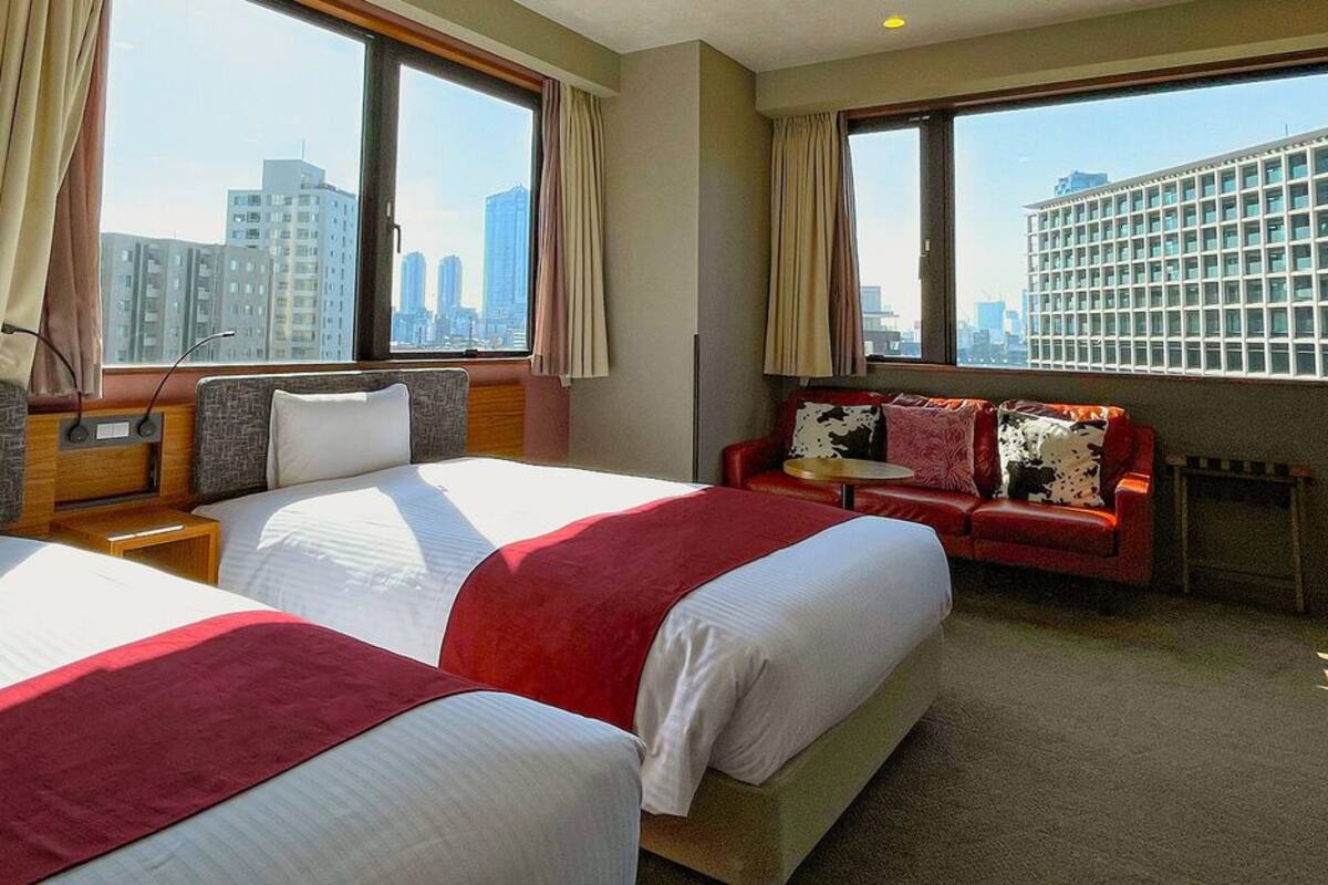 A hotel room with two beds, a sofa, a small table, and large windows with a city view.