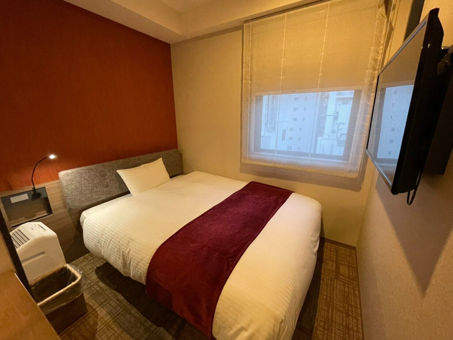 Quarto de hotel com uma cama, mesa de cabeceira, abajur, televisão e janela com persianas.