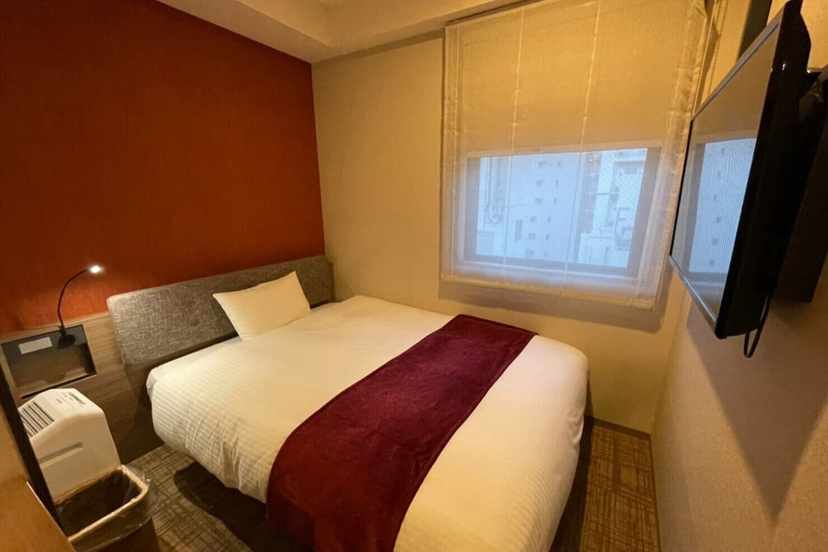 A hotel room with a bed, a nightstand, a lamp, a TV, and a window with blinds.