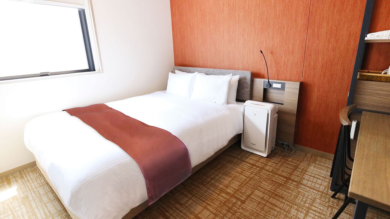 A hotel room with a bed, a desk, a chair, a heater, and a window.
