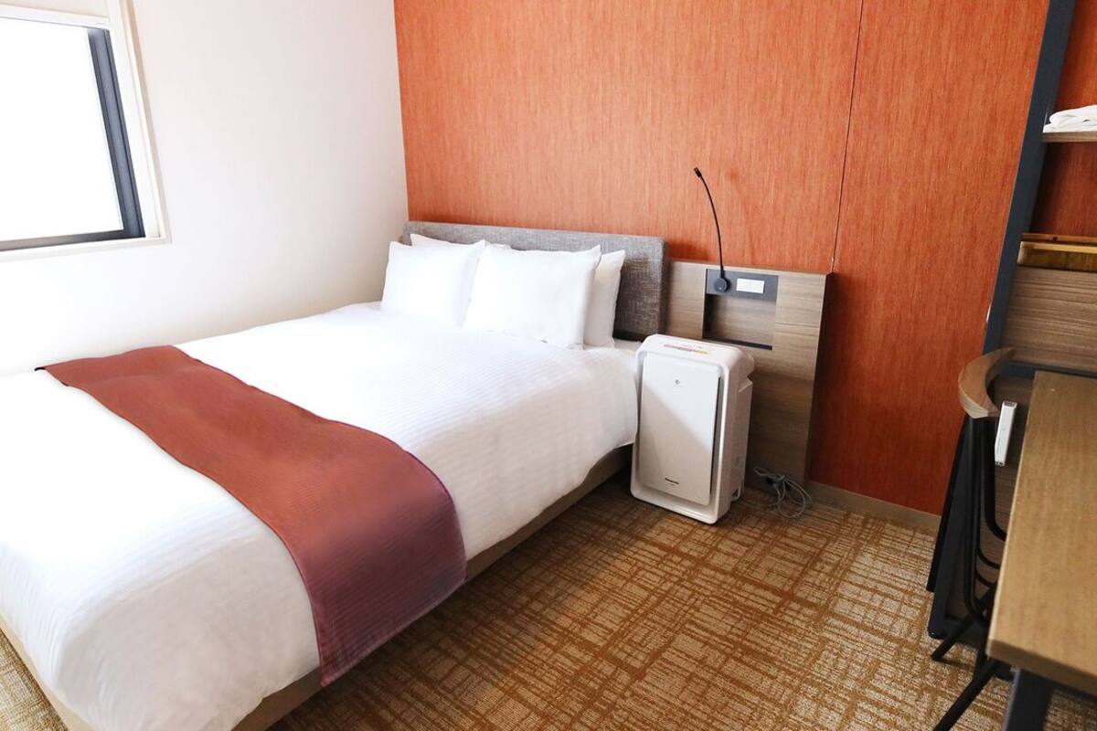 A hotel room with a bed, a desk, a chair, a heater, and a window.