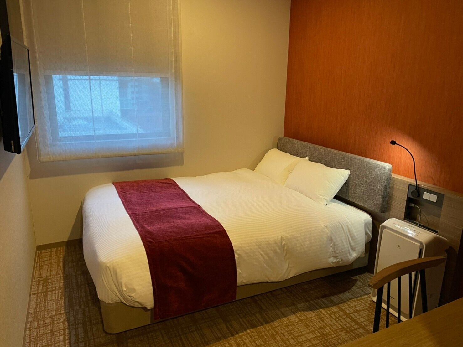 Quarto de hotel com uma cama, uma mesinha de cabeceira, uma luminária e uma janela com persianas.