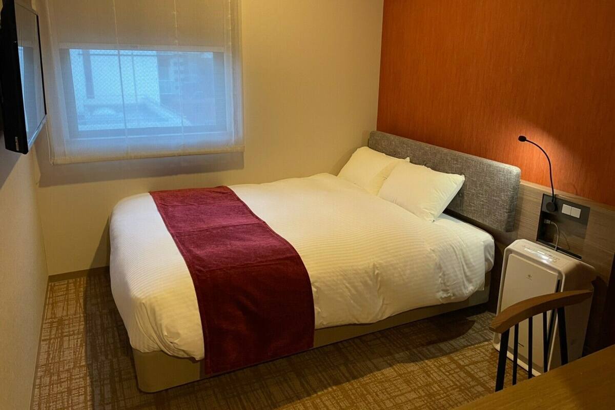 A hotel room with a bed, a small bedside table, a lamp, and a window with blinds.