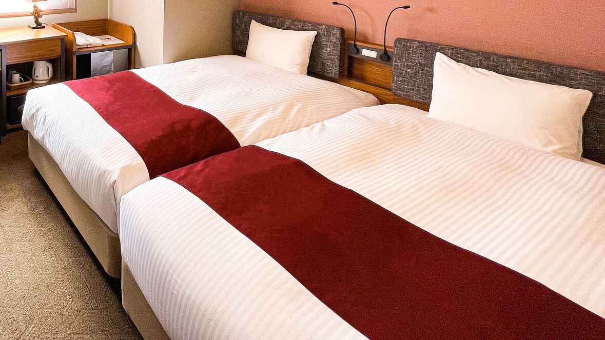 Two beds with white and red bedding, a wooden headboard, a nightstand with a lamp, and a small table with a kettle.
