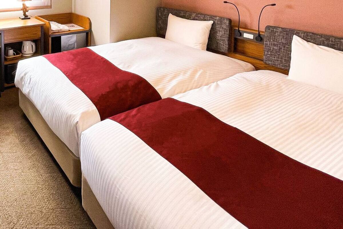 Two beds with white and red bedding, a wooden headboard, a nightstand with a lamp, and a small table with a kettle.
