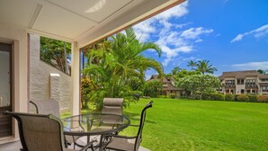 A glass-top table with four chairs, a stone wall, and a view of a well-maintained lawn and buildings in the background. - 1Br 1Ba Kahala Tropical Dream, Pool, Garden View 513 (Koloa)