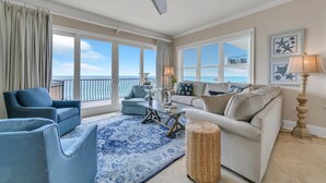 A spacious living room with a view of the ocean, featuring a ceiling fan, a large window with blinds, and a blue patterned rug. - Adagio A-205 - Amazing UPDATED Gulf Front Corner Unit! (Santa Rosa Beach)