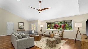 Ein geräumiges Wohnzimmer mit Deckenventilator, einem großen Fenster mit Blick auf Grünflächen und einer gemütlichen Sitzecke mit grauem Sofa und Poufs.