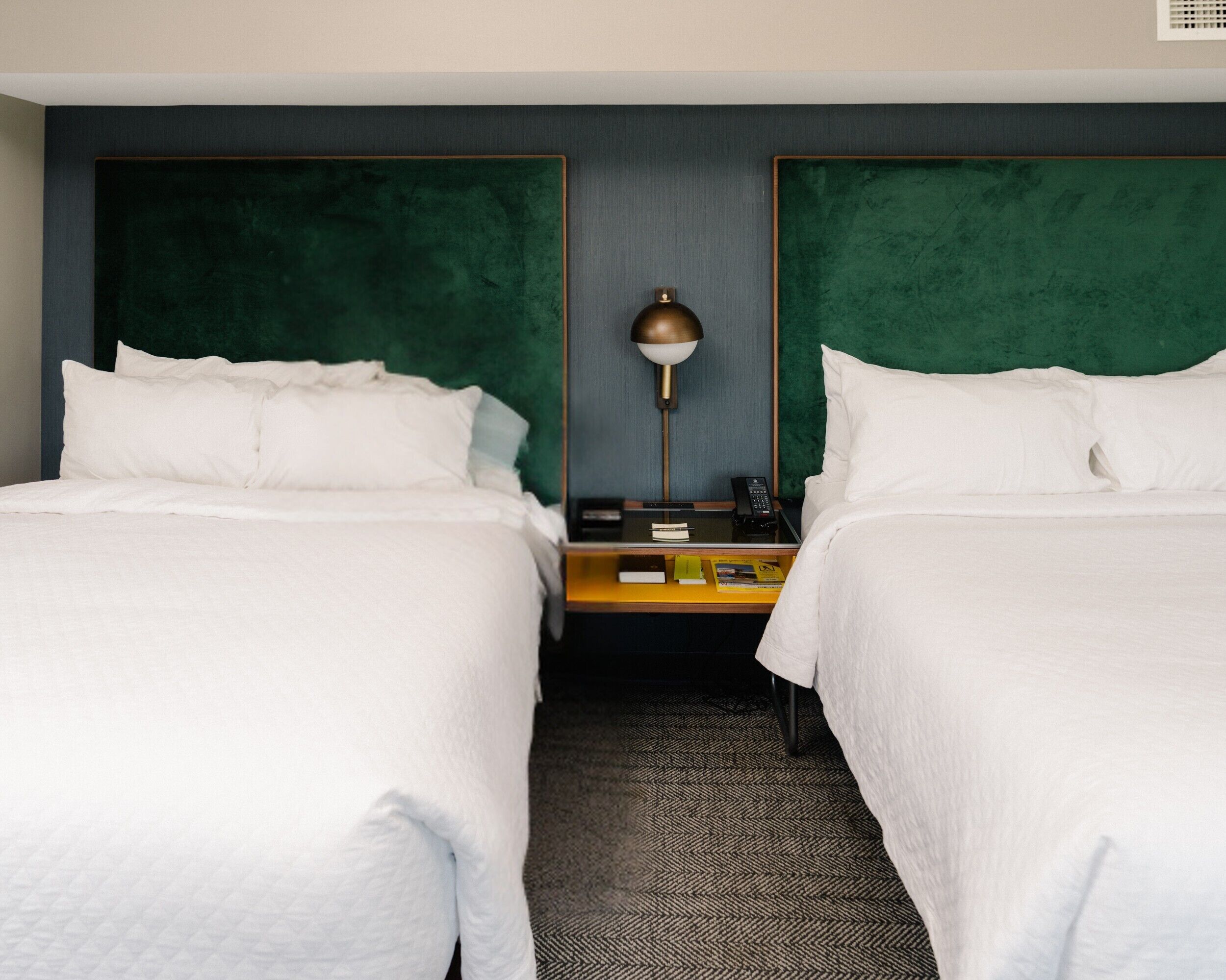 A hotel room with two beds, a nightstand with a lamp, and a small table with books and a phone.
