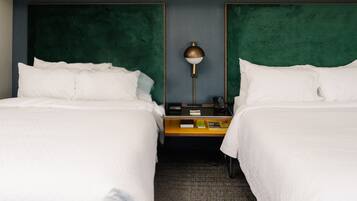 A hotel room with two beds, a nightstand with a lamp, and a small table with books and a phone.