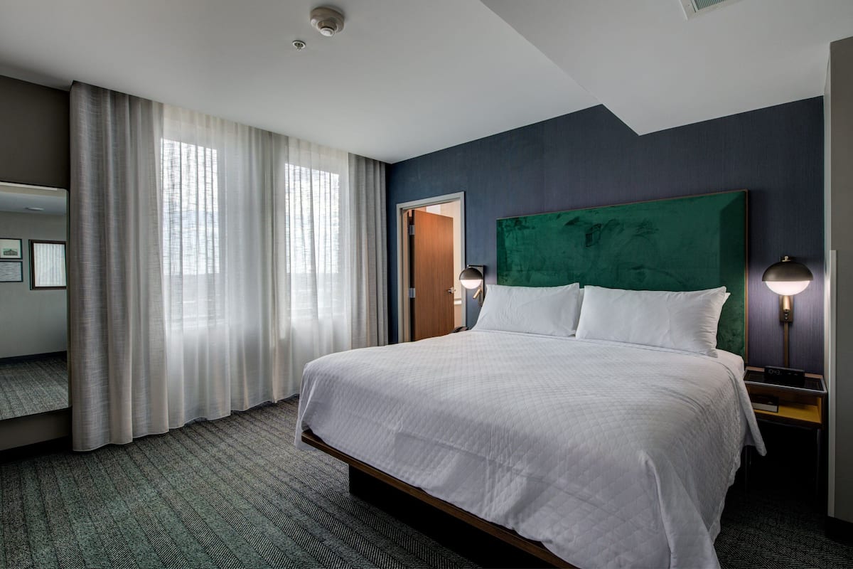 A hotel room with a large bed, a green headboard, a nightstand with a lamp, a window with curtains, and a door.