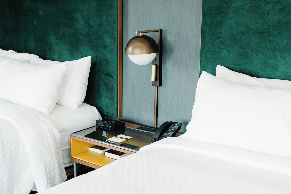 A hotel room with two beds, a nightstand with a lamp, a phone, and a small table with books and a notepad.