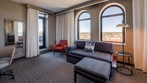 Quarto de hotel com sofá, poltrona e uma pequena mesa. Há uma janela grande com vista para a cidade.