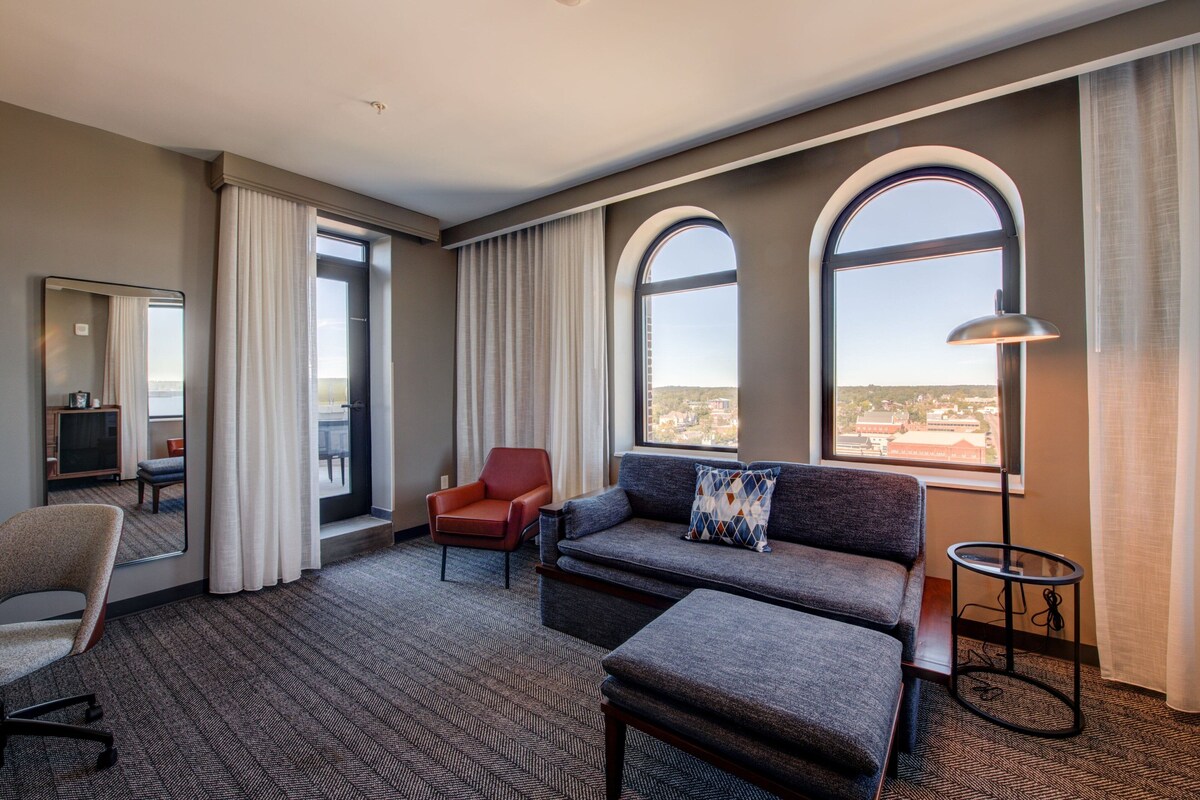 Habitación de hotel con sofá, sillón y una mesita. Amplia ventana con vistas a la ciudad.