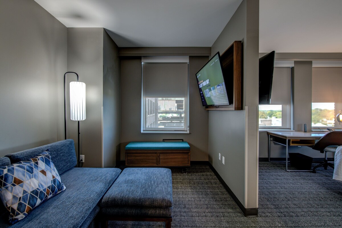 Habitación de hotel con sofá, banco, escritorio, televisión de pantalla plana y ventana con persianas.