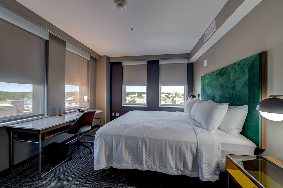 A hotel room with a large bed, a desk, and a chair. There are windows with blinds, a green headboard, and a lamp on the desk.
