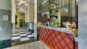 Um saguão de hotel com piso xadrez, uma recepção com espelho e um candelabro.