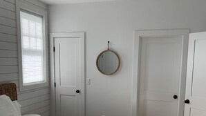 A bedroom with a large bed, two white doors, a round mirror, and a window with blinds. - Mid island oasis! 3-bedroom, 4 bath in the middle of “second downtown” (Nantucket)