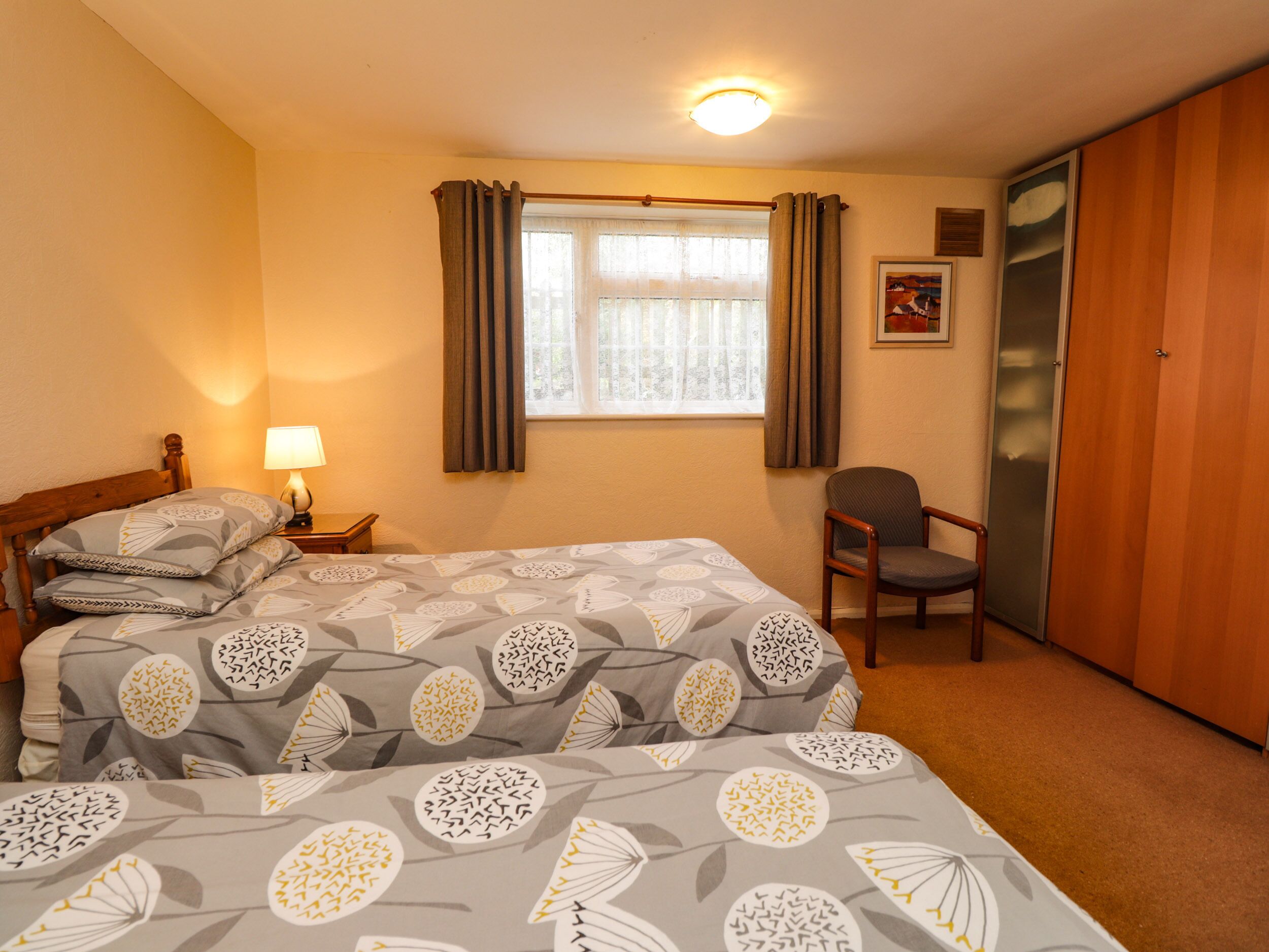 A hotel room with a bed, a chair, a nightstand with a lamp, a window with curtains, and a wardrobe.