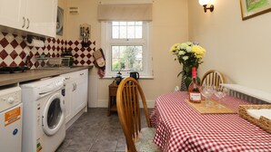 A compact kitchen with a washing machine, a dining table with a checkered tablecloth, and a window with a view of greenery. - Old Post Office (Ashford)