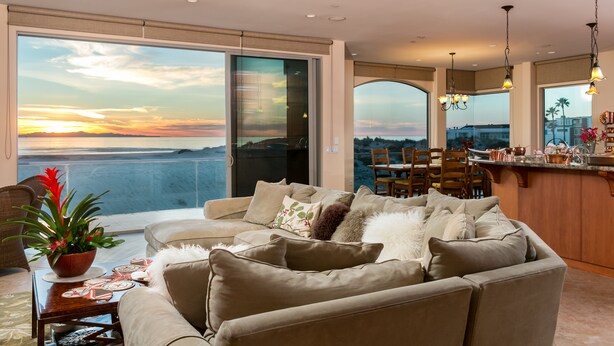 Uma sala ampla com um sofá grande, uma mesa de jantar e vista para o pôr do sol sobre a água.