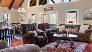 A spacious living room with a high ceiling, wooden beams, a chandelier, a fireplace, and a large window overlooking a forested area. - Halfback Ridge -Hot Tub-Magnificent Views-Motorcycle & Pet-Friendly-Fenced Yard
 (Maggie Valley)