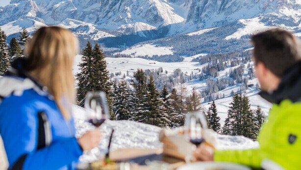 A group of people sitting at a wooden table with a view of snow-covered mountains in the background. - Binterhof vacation apartment GENZIANA (Trentino-Alto Adige)