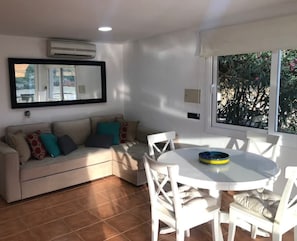 Uma sala de estar com um sofá branco, almofadas verde-água, uma mesa de jantar branca com cadeiras e uma janela com vista para áreas verdes.