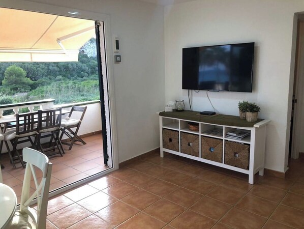 A room with a TV mounted on the wall, a balcony with a table and chairs, and a white console with storage drawers.