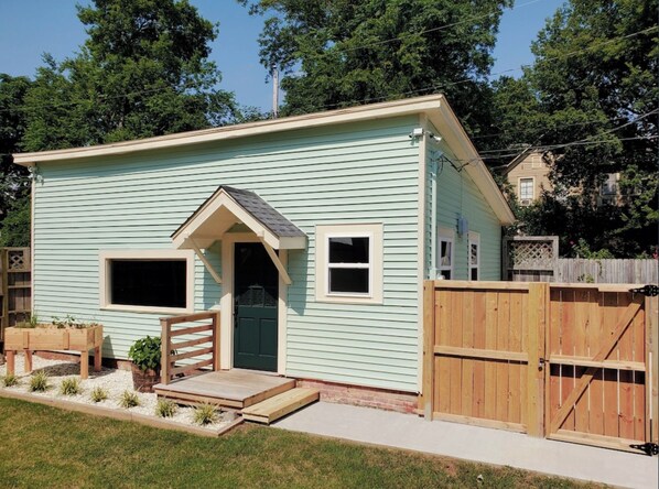 A small, single-story house with a light blue exterior, a front door, two windows, a small porch with steps, and a wooden fence. - The Carriage House in SoMa (Little Rock)