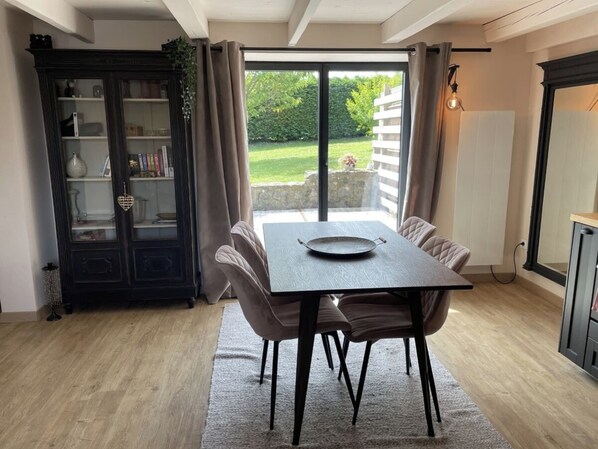 A dining area with a table and chairs, a dark wooden cabinet with glass doors, and a view of a garden through a sliding glass door.