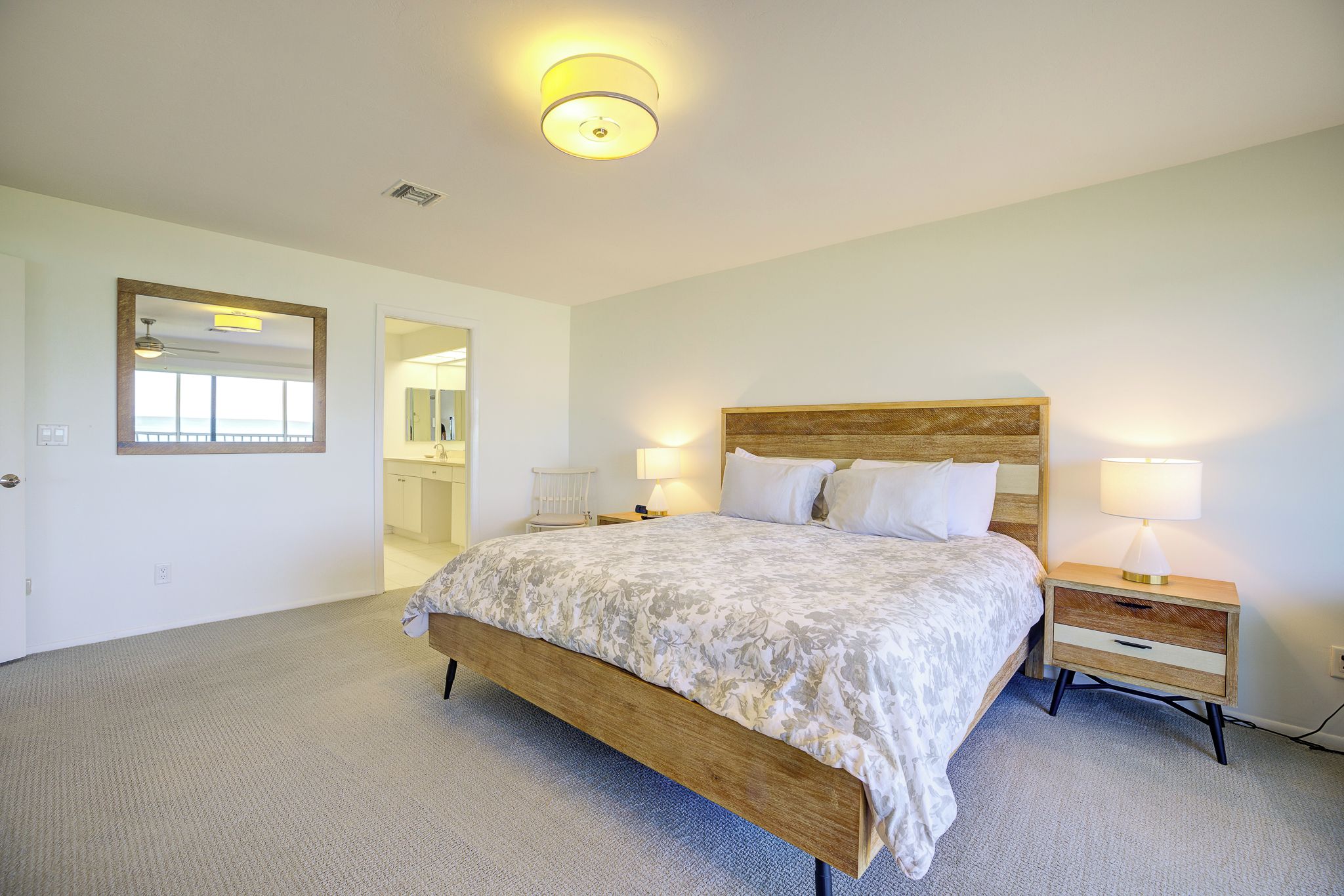 A bedroom with a wooden bed, two bedside tables with lamps, a mirror, and a door leading to a bathroom.