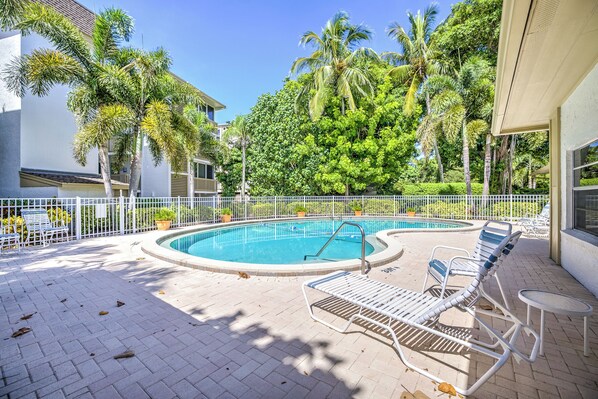 A swimming pool with lounge chairs and a fenced area, surrounded by palm trees and a building.