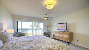 Habitación de hotel con una cama grande, ventilador de techo, televisión sobre una cómoda de madera y balcón con vista al océano.