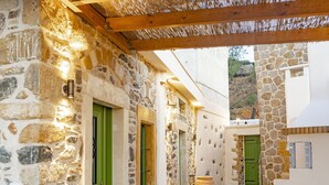 Un étroit couloir en pierre, avec des portes vertes, un grand vase et une pergola au-dessus.