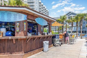 A wooden bar with a menu board listing drinks and cocktails, a trash can, and a person working behind the bar. - Beachfront Escape to Unwind & Take in the Ocean Air (Myrtle Beach)