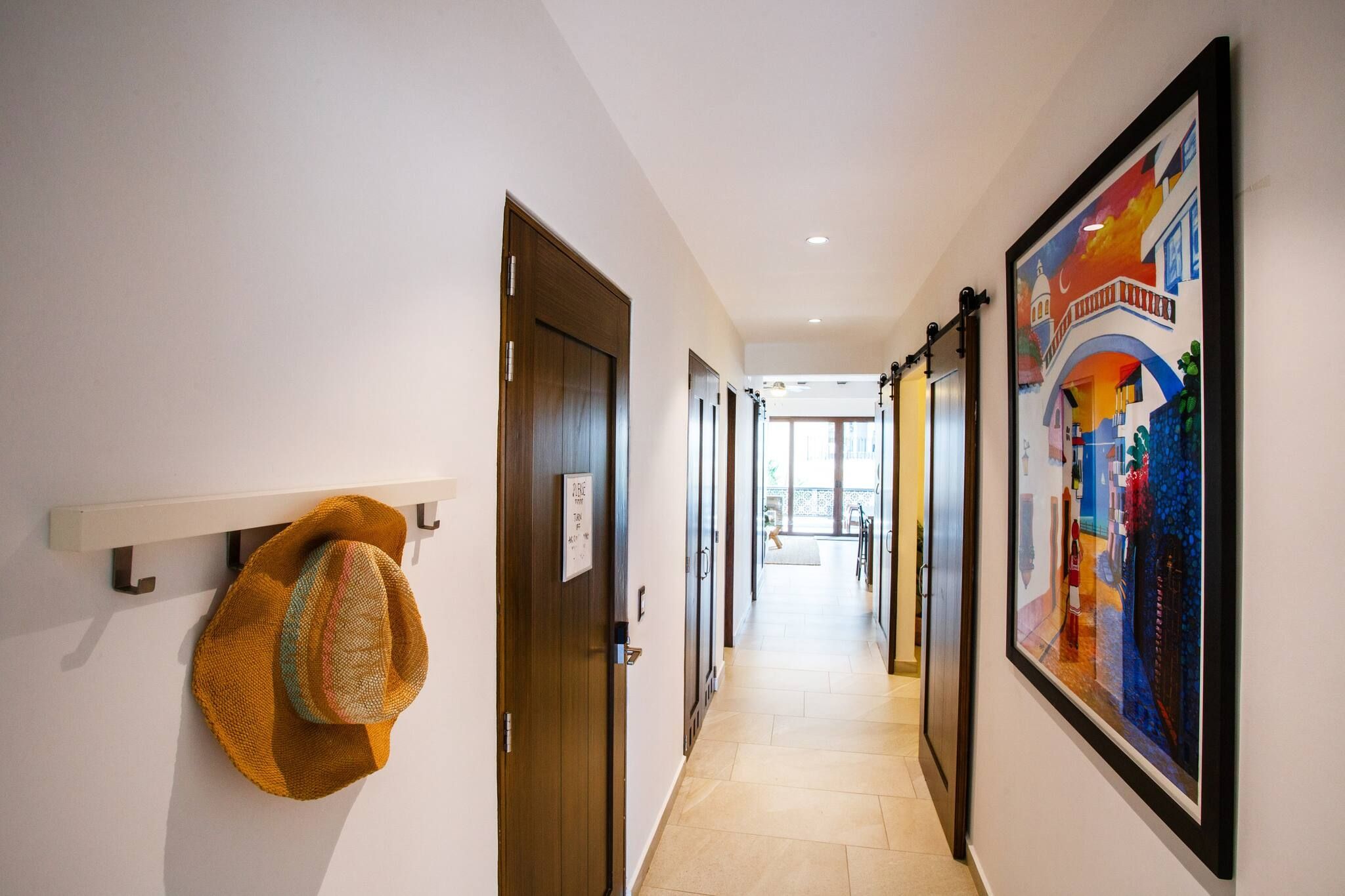 A hotel hallway with doors, a hat on a wall-mounted rack, and a colorful painting on the wall.