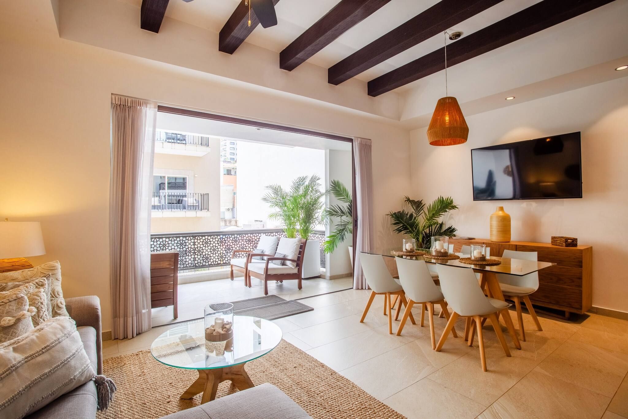 A modern living room with a glass coffee table, a sofa, a dining area with chairs, a TV mounted on the wall, and a balcony with plants.