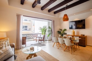 A modern living room with a glass coffee table, a sofa, a dining area with chairs, a TV mounted on the wall, and a balcony with plants. - Pool, 2 Balconies, Romantic Zone, Walk Everywhere (Puerto Vallarta)