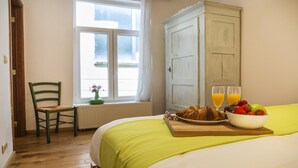 Un dormitorio con cama, una silla, un ventanal, un radiador y una mesa para el desayuno con cruasanes, fruta y zumo de naranja.