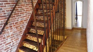 Una escalera de madera con barandillas de metal frente a una pared de ladrillo, que conduce a un pasillo con piso de madera y una puerta blanca.