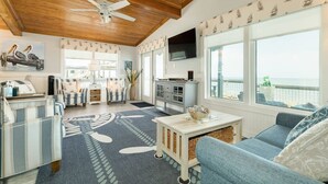 Un vaste espace de vie doté d’un plafond en bois, d’une grande fenêtre offrant une vue sur la mer, d’un téléviseur à écran plat fixé au mur et d’un coin salon confortable avec un canapé et des fauteuils.