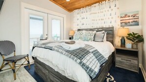 Une chambre avec un plafond en bois, un lit recouvert d’une couette à carreaux blancs et gris, une table de chevet avec une lampe, et une fenêtre avec des rideaux.