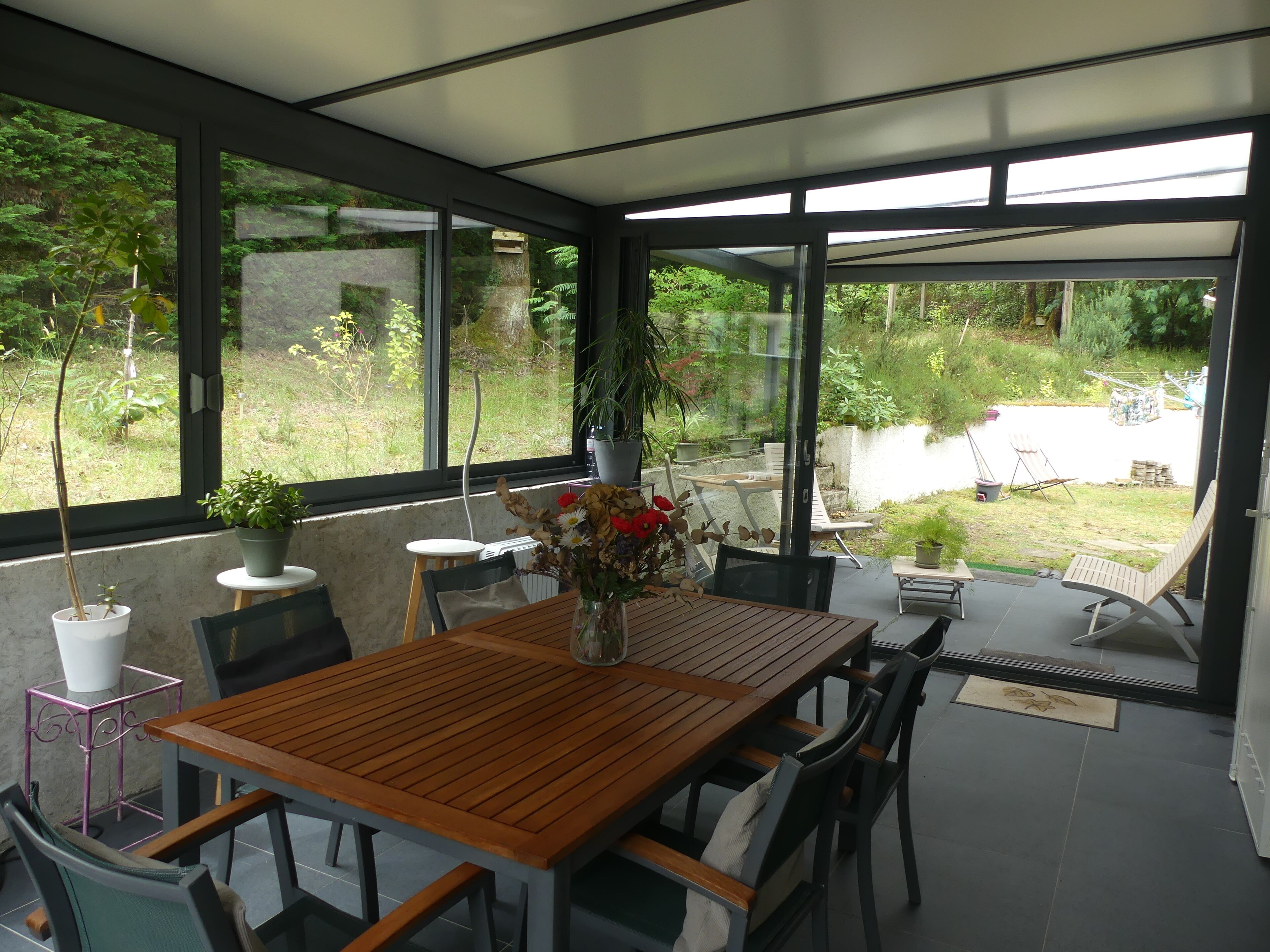 Un patio moderno con una mesa y sillas de madera, rodeado de amplios ventanales que dan a un exuberante jardín.