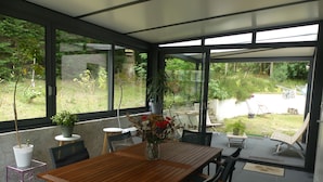 Un patio moderno con una mesa y sillas de madera, rodeado de amplios ventanales que dan a un exuberante jardín.