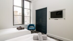 Habitación de hotel con dos camas, un ventanal grande, una silla azul, un armario oscuro y un televisor de pantalla plana montado en la pared.