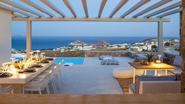 A terrace with a pool, dining area, and seating with a view of the sea and buildings.