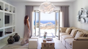 A modern living room with a sofa, a coffee table, and a TV. There is a view of the sea through large windows.