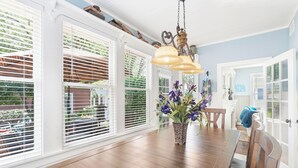 Un coin repas avec une table en bois, des stores blancs, un luminaire suspendu et un vase rempli de fleurs.