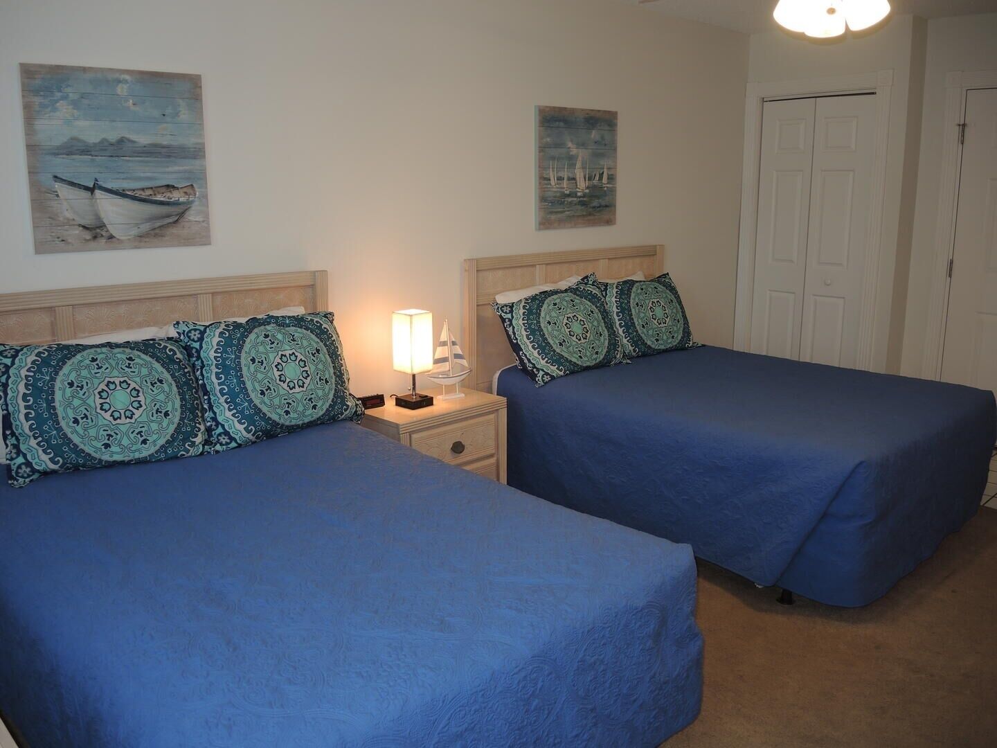 A room with two beds, each with matching headboards and blue bedding. There are two paintings on the walls, one depicting boats and the other sailboats. A nightstand with a lamp is between the beds.